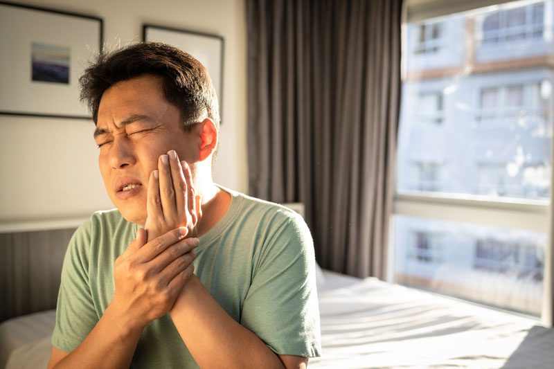 A person holds their cheek in pain, eyes closed, in a brightly lit bedroom with framed art and large windows.