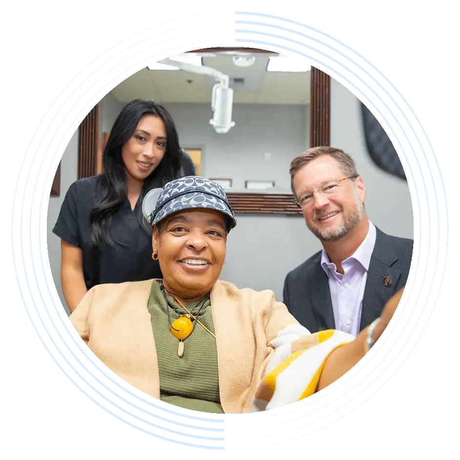A person in a cap smiles and holds a phone for a selfie, with a man in a suit and a woman in scrubs standing behind, inside a medical office.