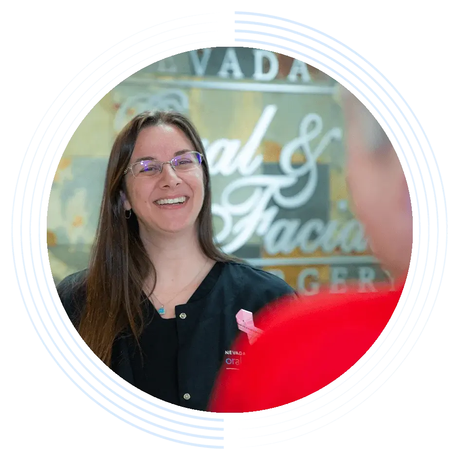 A woman wearing glasses and a black jacket smiles in conversation. "Nevada Oral & Facial Surgery" is partially visible on the wall behind her, indicating a professional setting.
