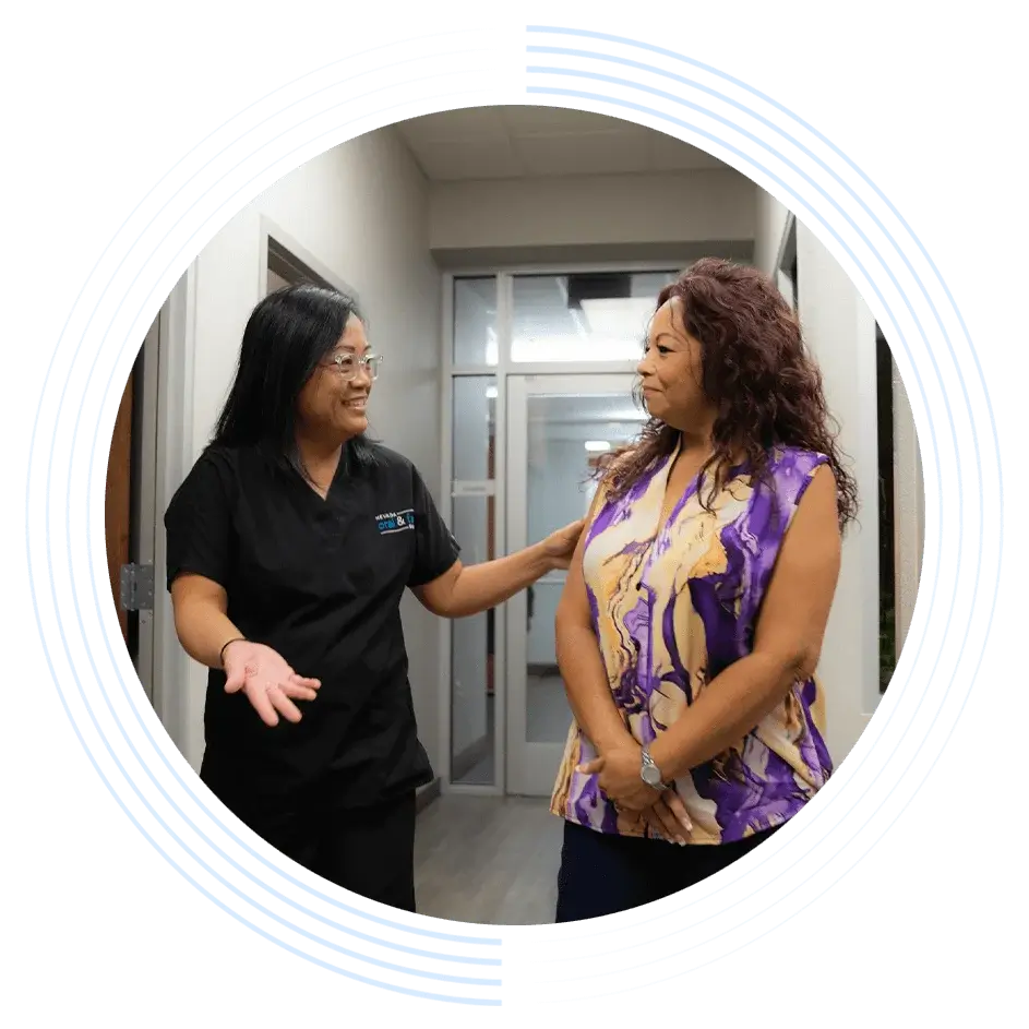 A woman in black scrubs talks to another woman in a colorful blouse in a hallway with gray walls and glass doors. The woman in scrubs is smiling and gesturing.