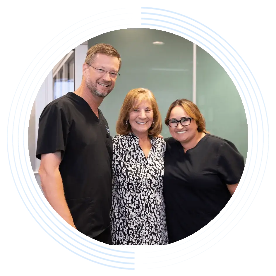 Three people smile, standing close together in a professional environment, framed by a decorative circular pattern. The setting features light-colored walls and large windows in the background.