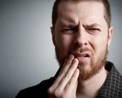 A bearded man touches his cheek with a pained expression, likely indicating discomfort. He is in a neutral, dimly lit environment, suggesting a focus on his facial expression.