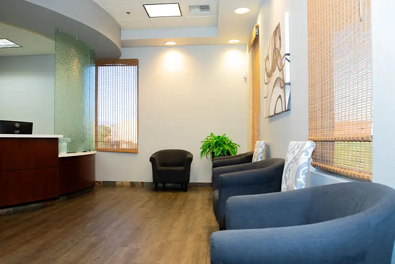 Chairs line a waiting area with wood flooring; a reception desk is on the left, artwork on the wall, and a plant decorates the space.