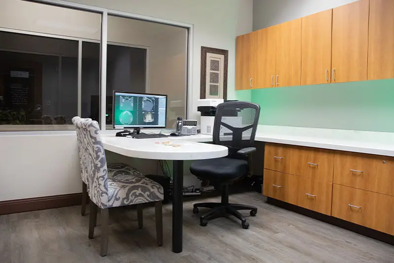 A computer displays scans on a round, white desk with ornate and black office chairs. Context: modern office with wooden cabinets, phone, and windowed wall; bright, organized setting.
