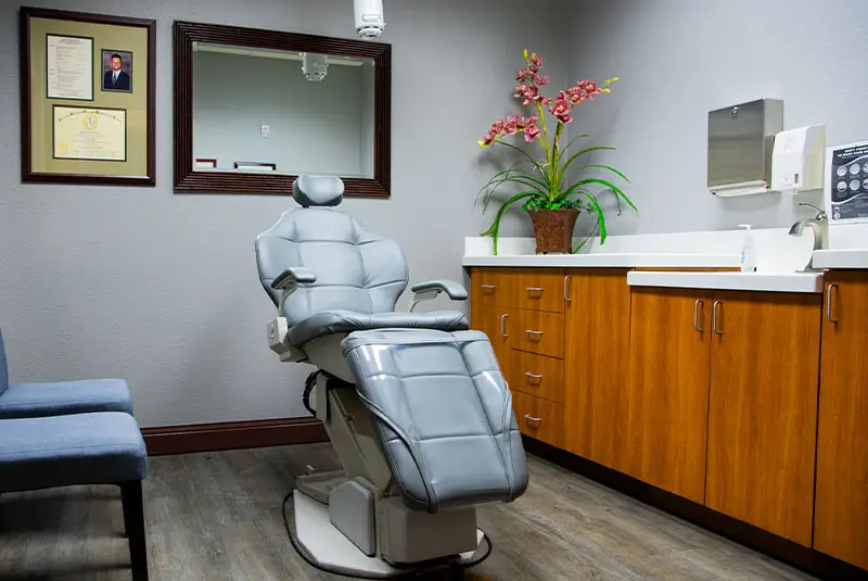 A gray dental chair sits centrally in a room with wooden cabinets, framed certificates, and a plant on the counter. A mirror and a wall-mounted paper towel dispenser are visible.