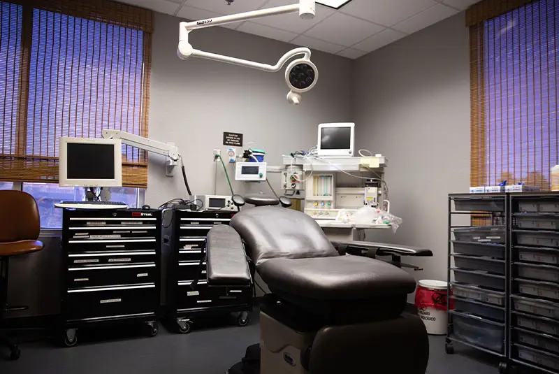 A medical examination chair is positioned in a clinical room, surrounded by monitoring equipment, computer screens, medical cabinets, and surgical lights. Walls have windows with woven blinds.