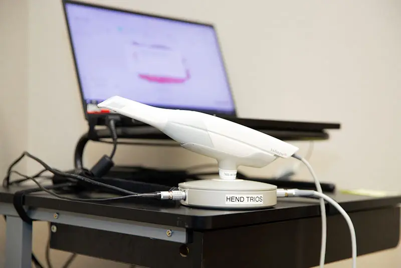A dental scanning device labeled "HEND TRIOS" rests on a table, connected by cables. It is next to a laptop displaying a blurry screen in an office setting.