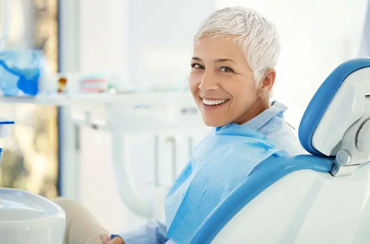 A person with short gray hair smiles while sitting in a dental chair, wearing a blue bib. Dental equipment and a bright, clinical environment surround them.