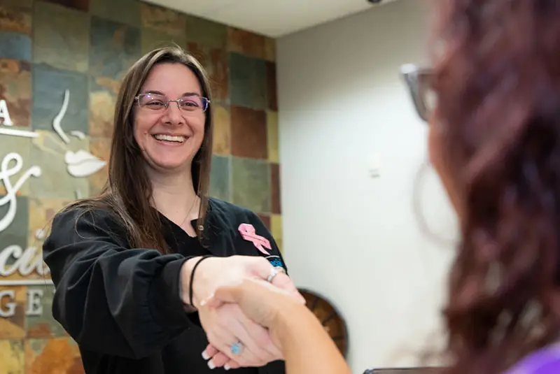 A smiling woman wearing glasses and a black shirt with a pink ribbon shakes hands with another person. They are in a room with tiled walls, partially displaying signage.
