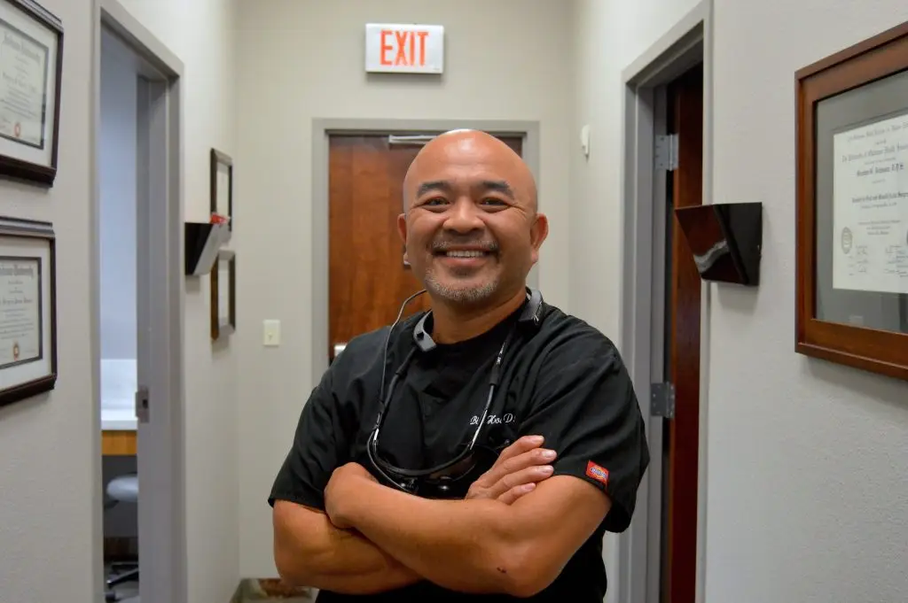 A person in a black uniform stands smiling with arms crossed in a hallway lined with framed certificates and an exit sign above a wooden door at the end.