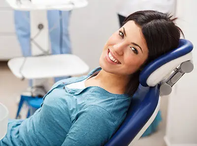 A woman in a blue shirt is smiling while reclining in a dental chair. The background shows dental equipment and a person wearing medical scrubs.