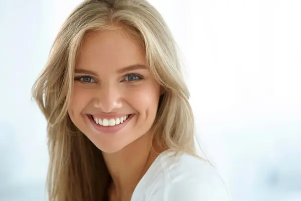 A person with long blonde hair smiles warmly, wearing a white top in a brightly-lit, soft-focus setting.
