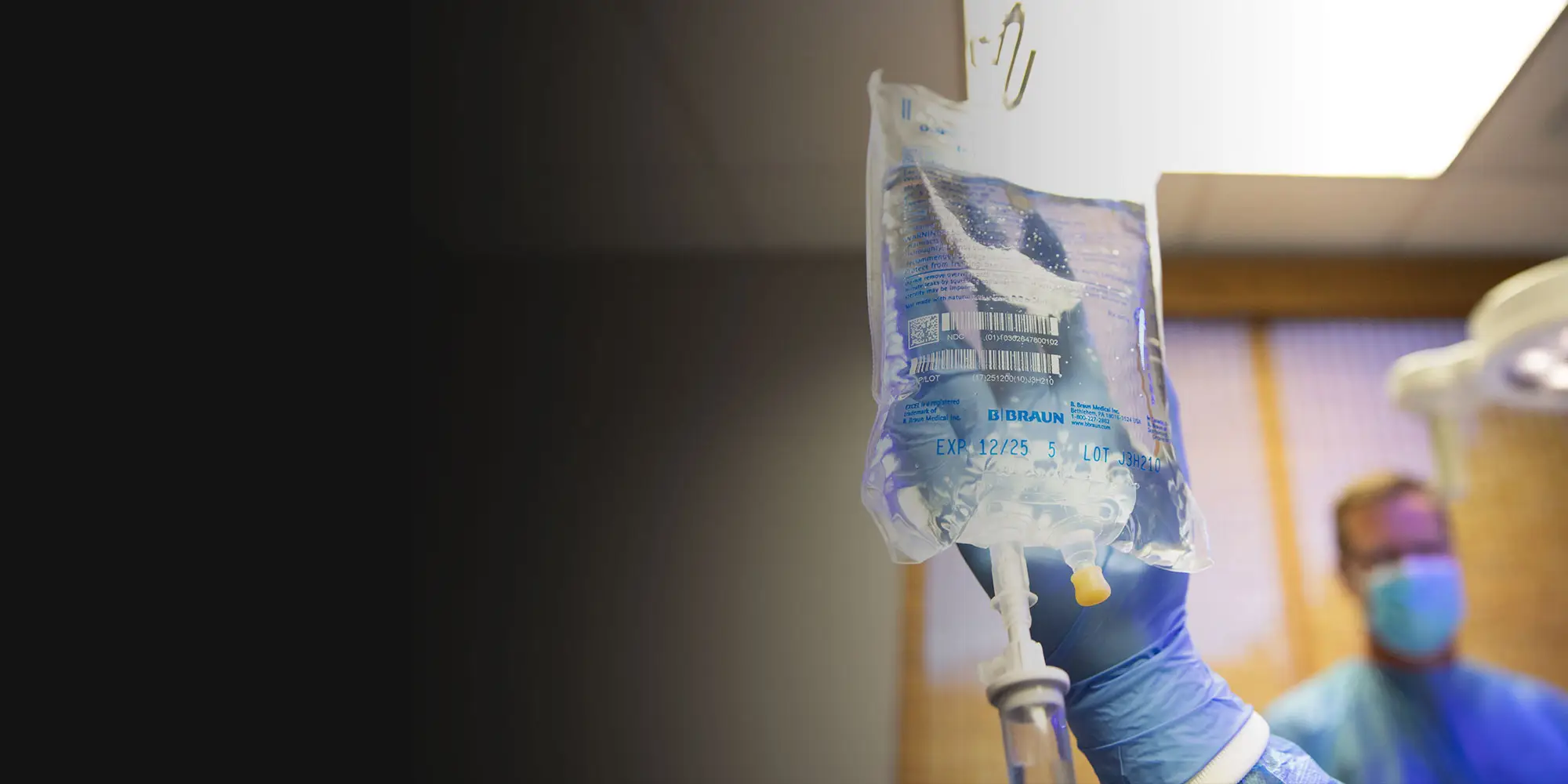 A gloved hand adjusts an intravenous fluid bag labeled "EXP 12/25" in a brightly lit medical room. A healthcare professional in a mask and gown stands blurred in the background.