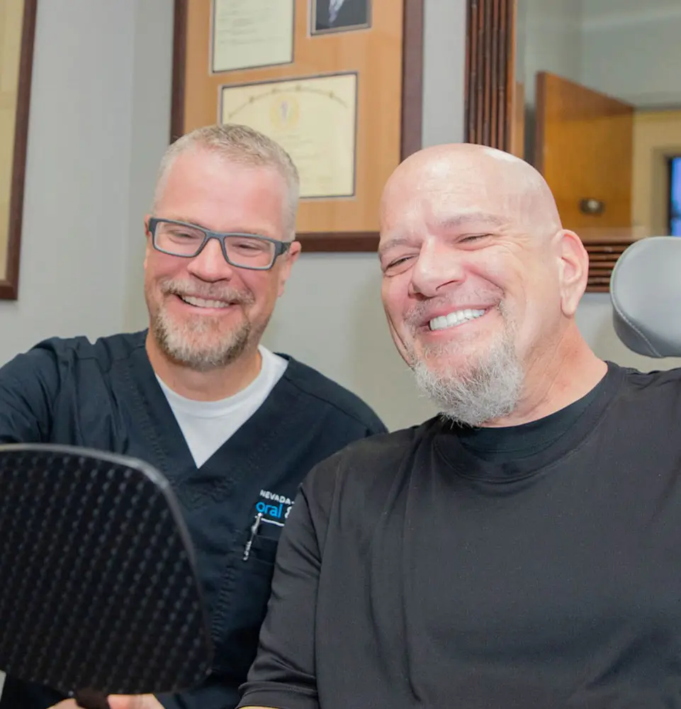 Two men sit smiling, one holds a mirror. Certificates and framed photos hang on the wall in a professional office setting.