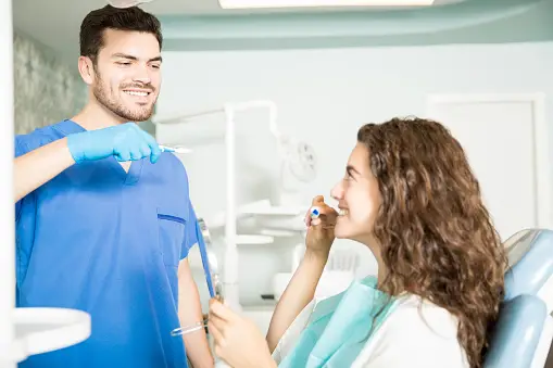 Dentist in blue scrubs demonstrates proper brushing technique to a seated patient holding a toothbrush, in a dental office with equipment visible in the background.