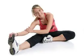A person in athletic clothing stretches by sitting with one leg extended and one bent, reaching for their foot, on a white background.