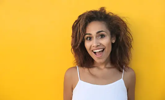 A person is smiling widely, standing against a bright yellow background. They are wearing a white sleeveless top and have curly hair.