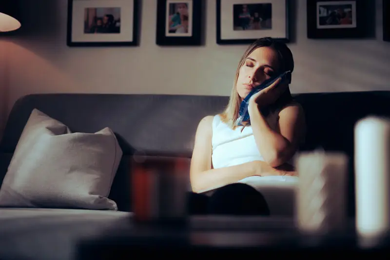 A person sits on a couch, eyes closed, holding an ice pack to their cheek. Behind, framed photos hang on the wall, and a soft lamp light illuminates the scene.