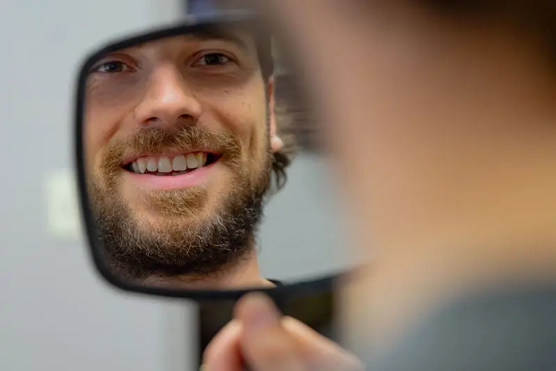 A person smiles at their reflection in a small handheld mirror, revealing a beard and teeth. The mirror is held indoors, partially reflecting a blurred background.