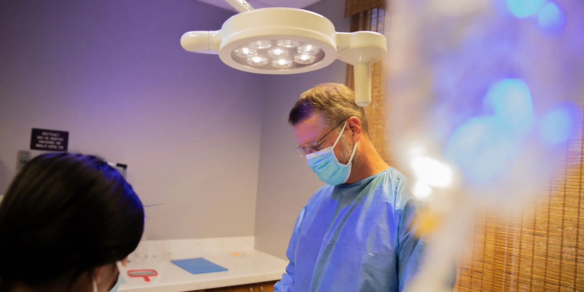 A medical professional in scrubs and mask focuses under surgical lights; nearby, another masked person assists in a clinical room. A wall-mounted sign reads: "RESTROOM ON SECOND FLOOR."