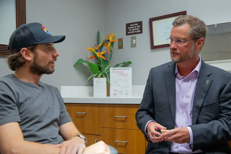 A seated man in a suit converses with another man in casual attire in an office setting, with a desk, tropical plant, framed documents, and a “Cover Your Cough” poster nearby.