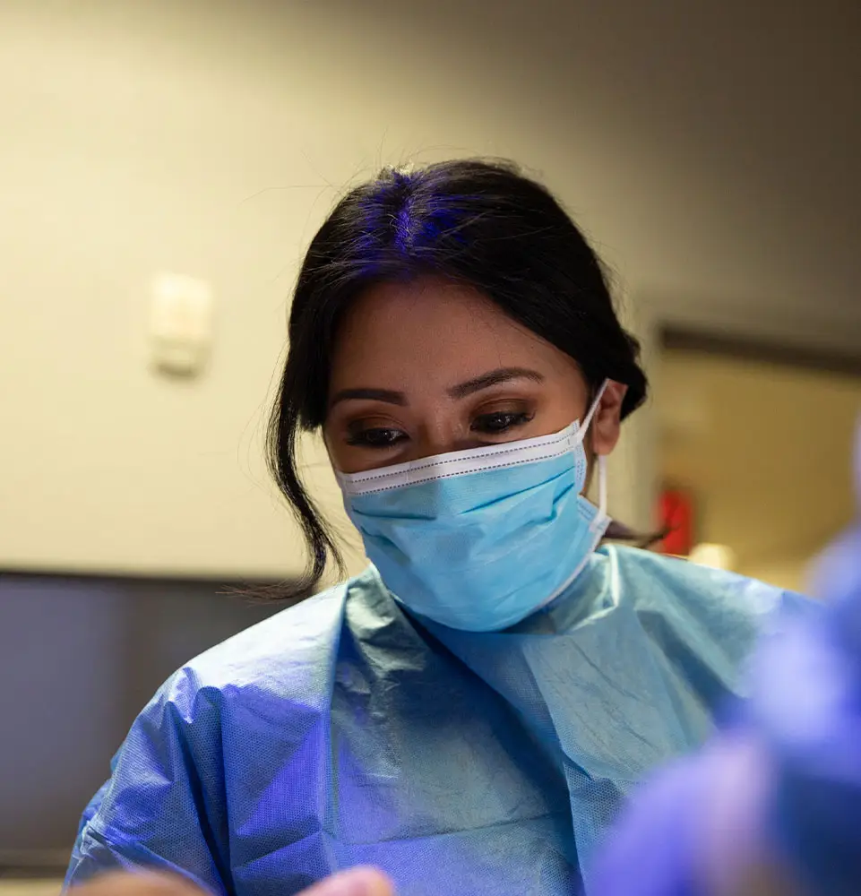 A person wearing a blue surgical mask and gown looks downward, focusing intently. The surrounding environment appears to be a clinical or medical setting with blurred equipment.