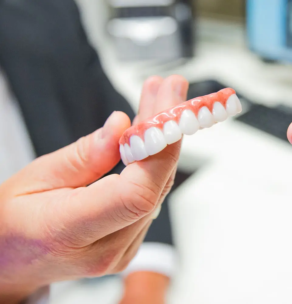 A person holds a set of upper dentures in a bright, clinical setting, with a computer and paperwork visible in the background.