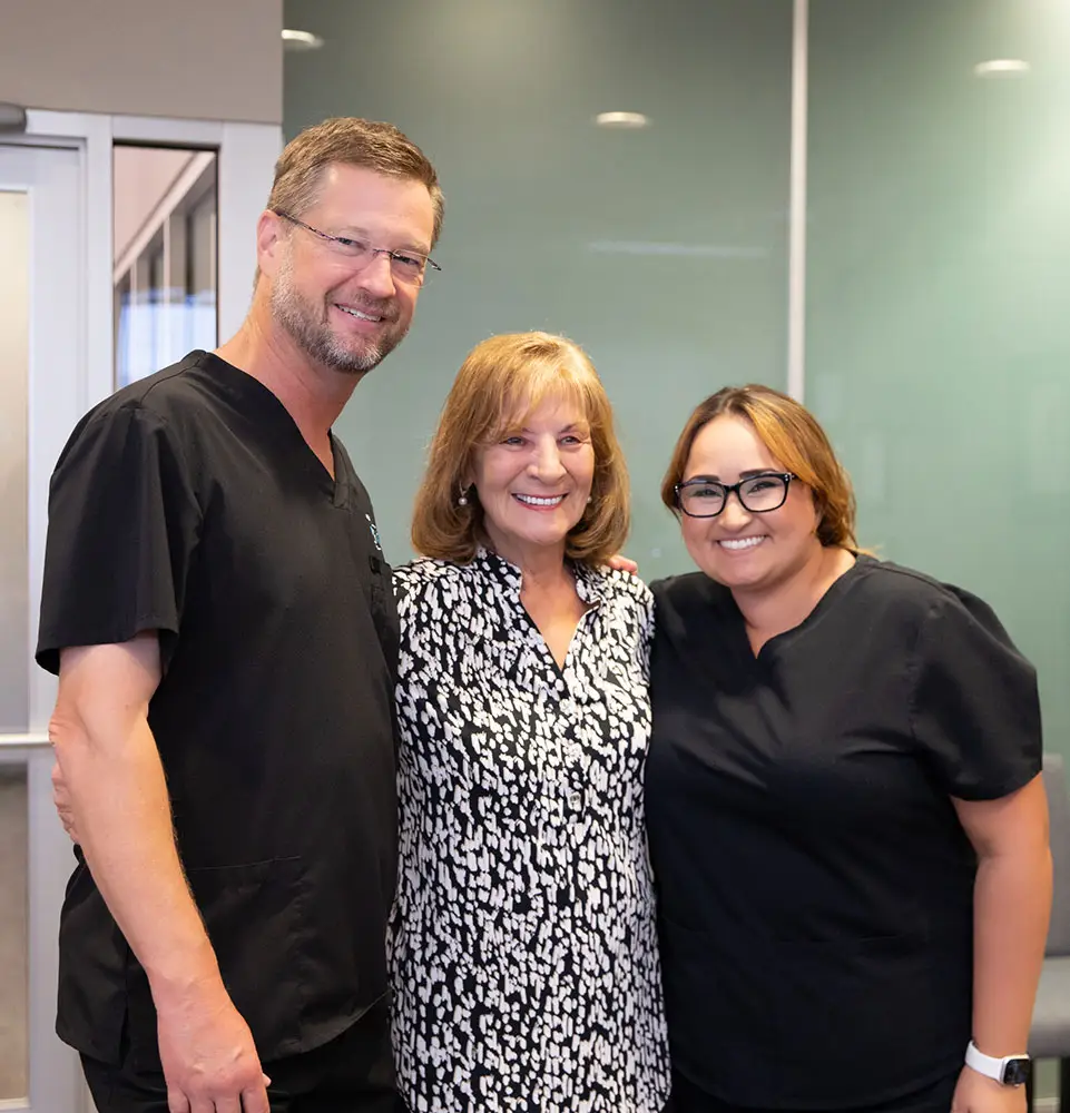 Three people smiling, standing together. Two wear black scrubs, suggesting medical professionals, and the other wears a patterned blouse. They're in an indoor setting, possibly a clinic or office. No text present.