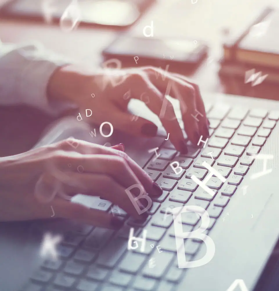 Hands type on a white keyboard with floating letters, suggesting digital writing or communication, on a desk with electronic devices and books in soft focus. No text present.