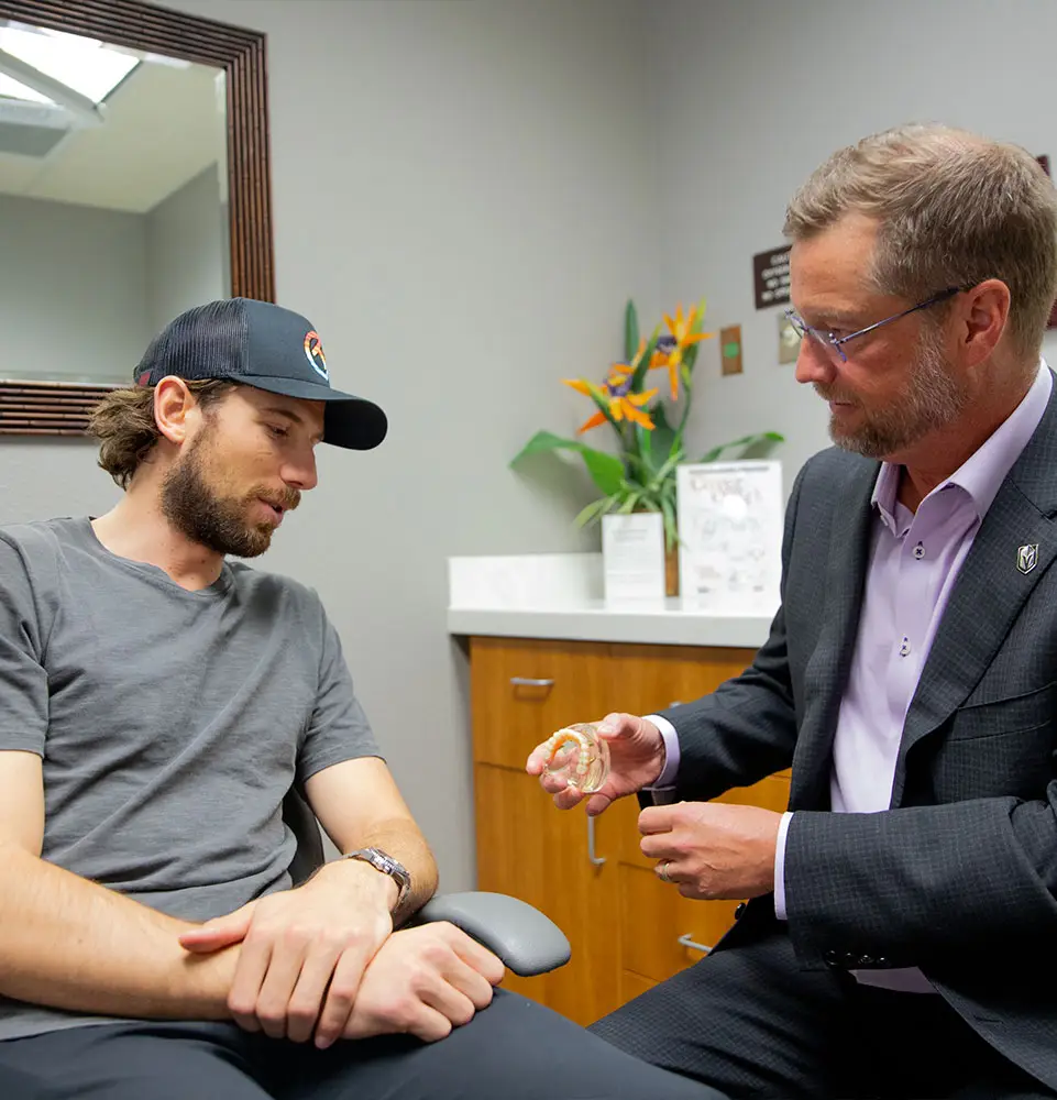 A man sitting in a chair listens to another man in a suit holding a dental model, in a clinical office with a plant and documents on a counter.