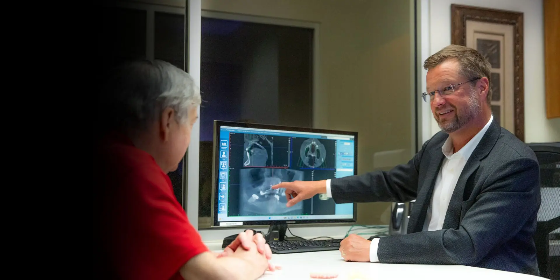 A man in a suit points at dental X-rays displayed on a computer monitor, sitting with another person at a desk in an office setting.