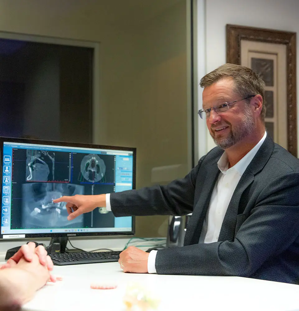 A man in a suit points at dental X-rays displayed on a computer monitor, sitting with another person at a desk in an office setting.