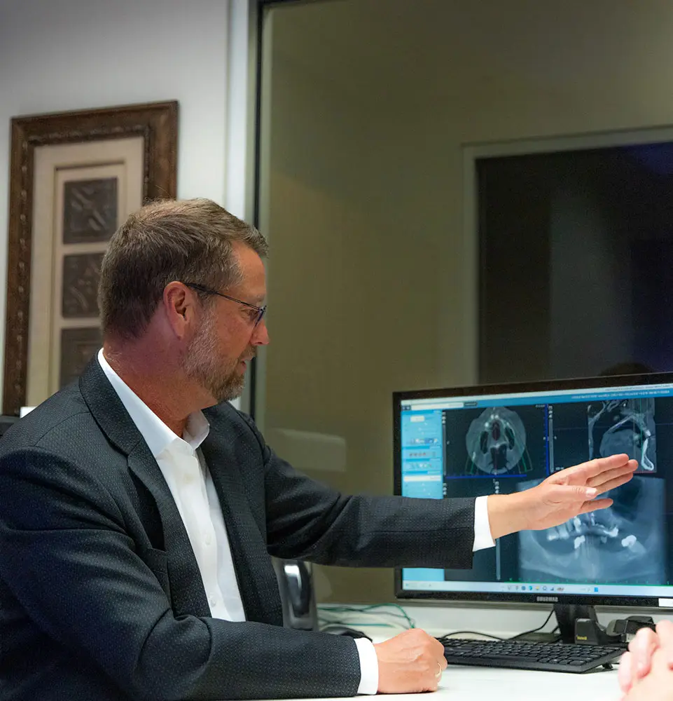 A person in a suit points at medical scans on a computer monitor in an office setting, discussing or analyzing the images.