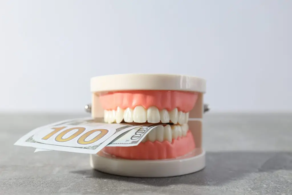 Dentures grip a $100 bill, positioned on a gray surface in a bright, minimalist setting.