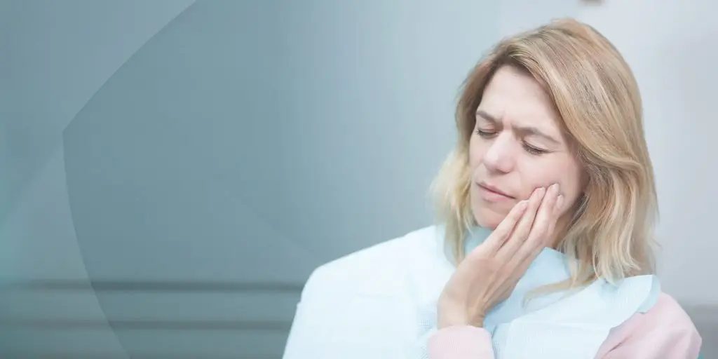 Middle-aged woman wincing and holding her cheek in pain while wearing a dental bib against a soft blue background.