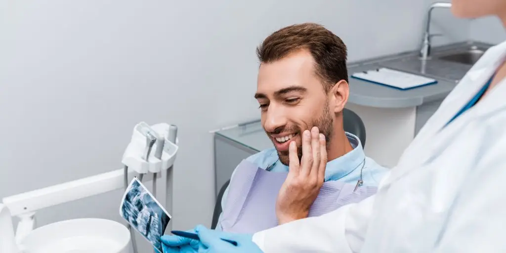 A patient sits in a dental chair, smiling and touching his cheek, while a dentist shows him a dental X-ray. The setting includes dental equipment and a counter with a clipboard.