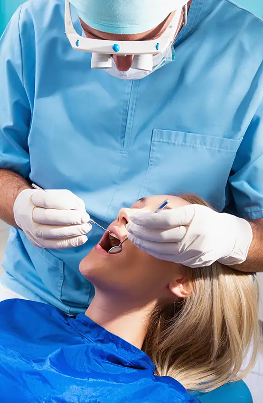 A dentist examines a patient's mouth using dental tools. The dentist wears blue scrubs and gloves, while the patient lies back, covered with a protective blue cloth.