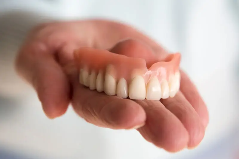 A set of upper dentures rests in an open palm, with a blurred light background, highlighting the detailed teeth and gum design.
