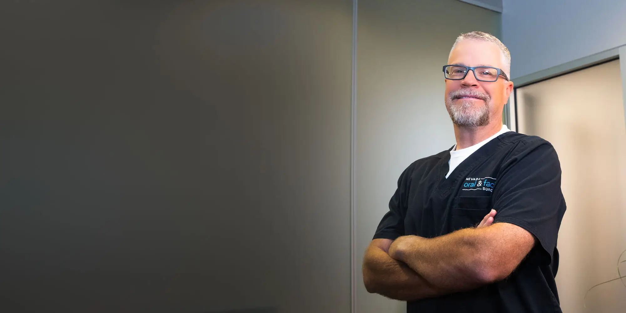 A man with glasses and a beard stands confidently with arms crossed, wearing a black shirt labeled "Nevada Oral & Facial Surgery," in a well-lit room with plain walls.