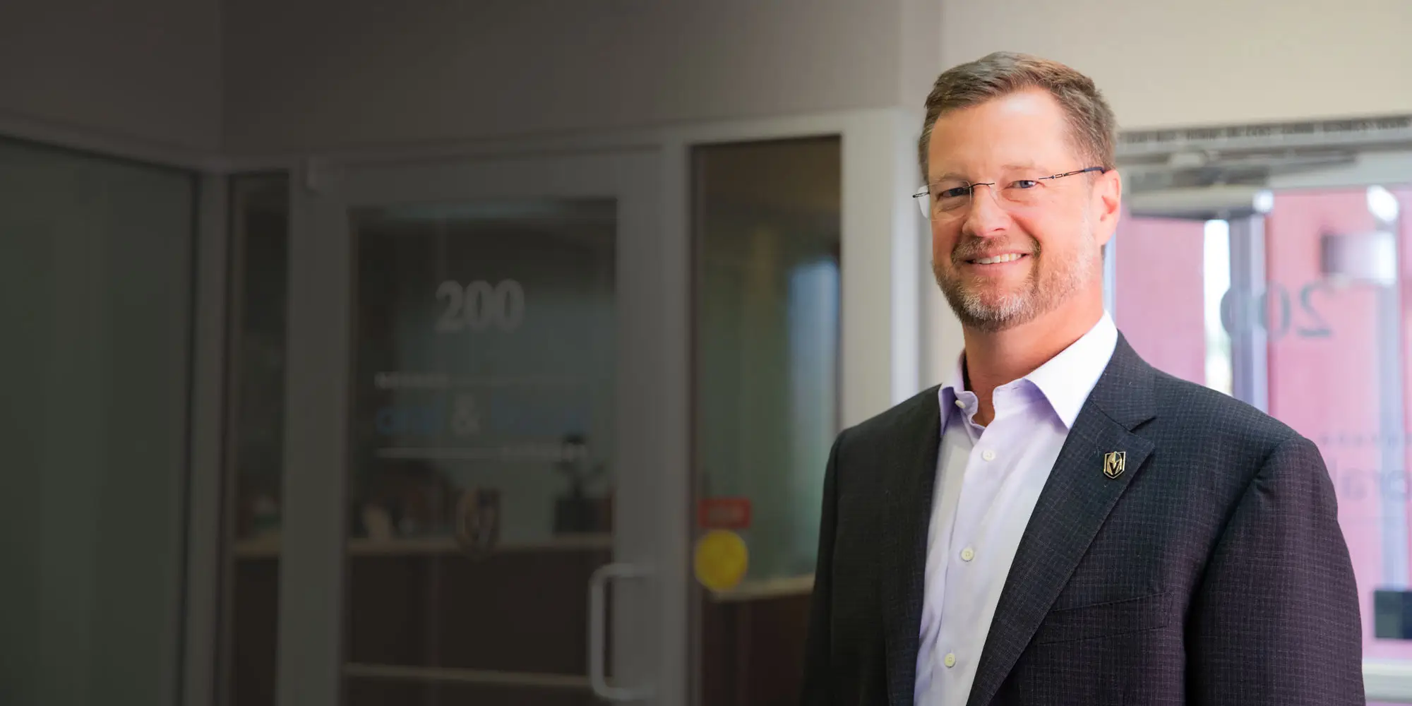A person in a suit stands smiling in an office lobby, near glass doors labeled "200" and "CNR &" with blurred interior elements in the background.