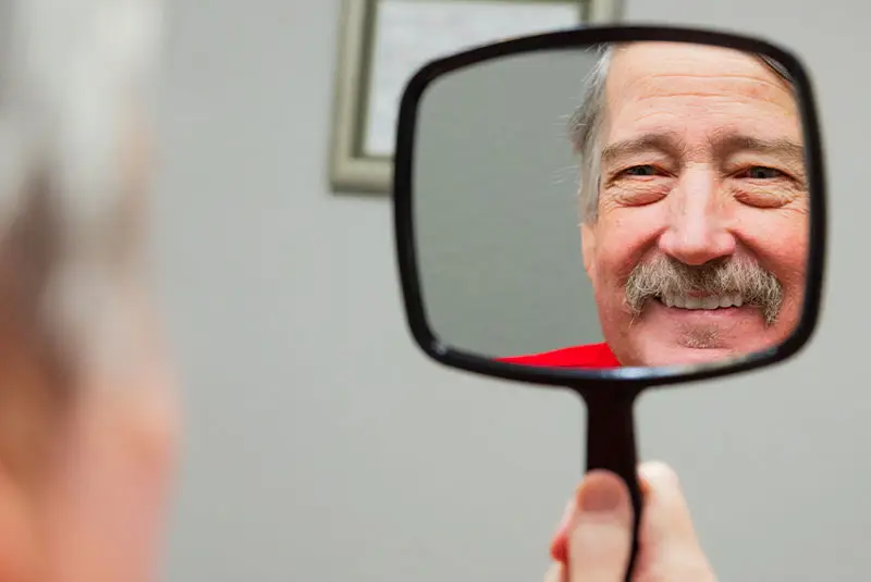 A person holds a handheld mirror reflecting their smiling face, framed by a mustache, in a room with a blurred picture frame on the wall behind them.