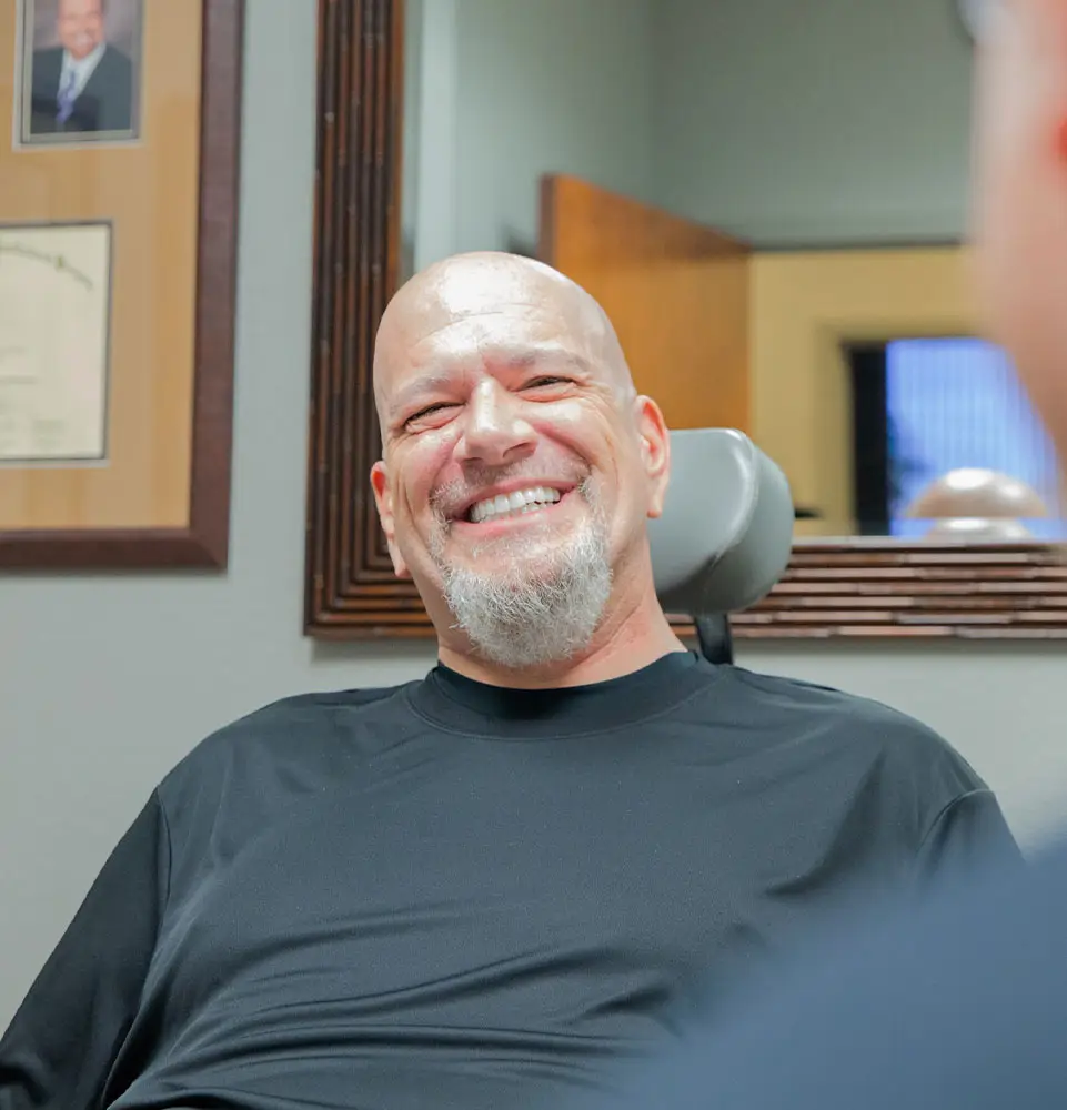 A man in a suit laughs while holding a dental model in an office with wooden cabinets and a framed picture on the wall.