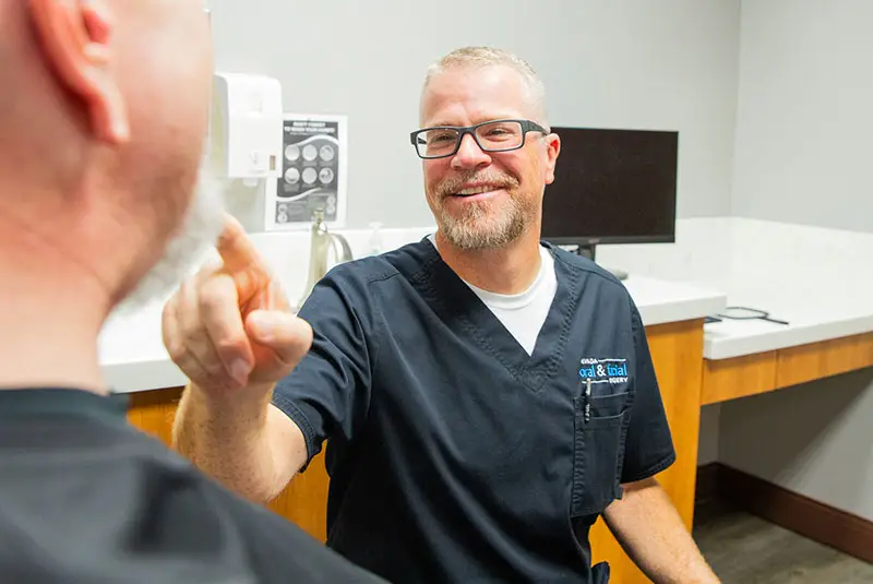 A smiling man in medical scrubs gestures warmly towards another person in a clinical room with a counter and computer nearby, conveying a welcoming, professional atmosphere.