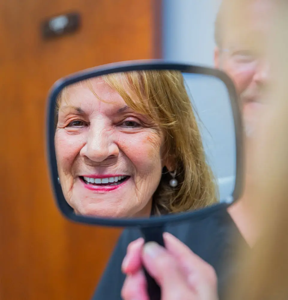 A woman smiles brightly, reflected in a handheld mirror, with a blurred indoor setting in the background.