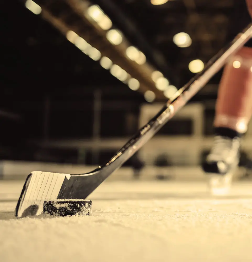 A hockey stick pushes a puck across the ice in an indoor rink. Nearby, a skater's legs and blurred lights overhead suggest motion and action during a game or practice.