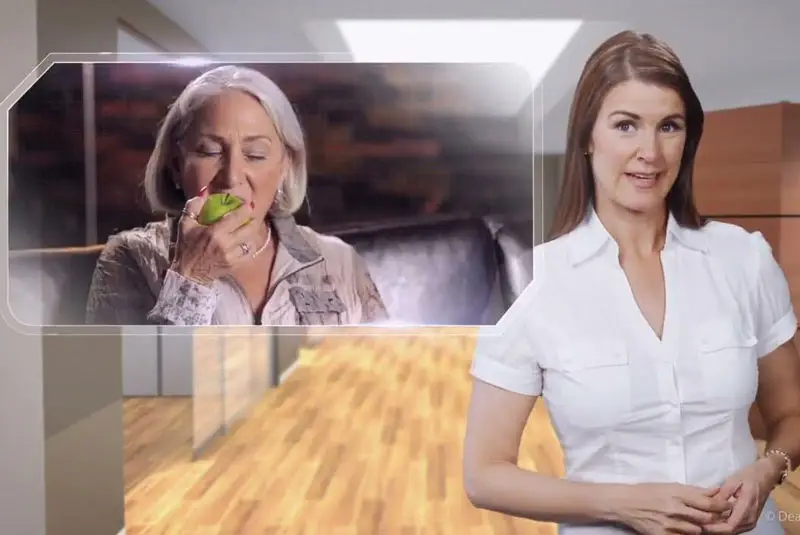 A woman wearing a white blouse stands in a modern room, gesturing towards a projected image of an elderly woman eating a green apple on a couch.