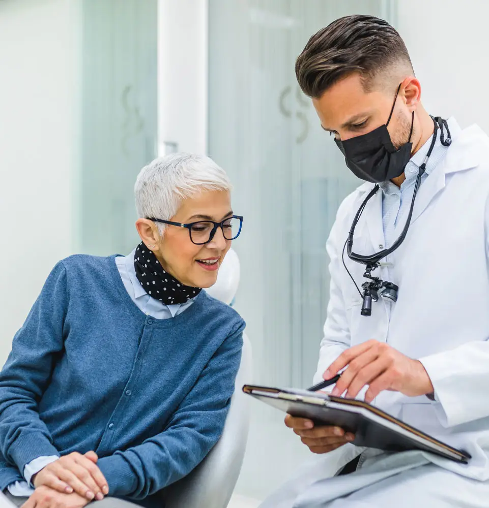 A dentist wearing a mask shows a clipboard to a smiling woman sitting in a dental chair; they are in a modern dental office.