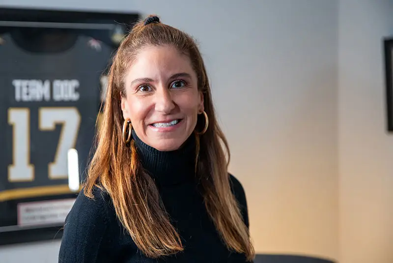 A person in a black turtleneck smiles indoors, with framed jersey displaying "TEAM DDC 17" on the wall behind them.