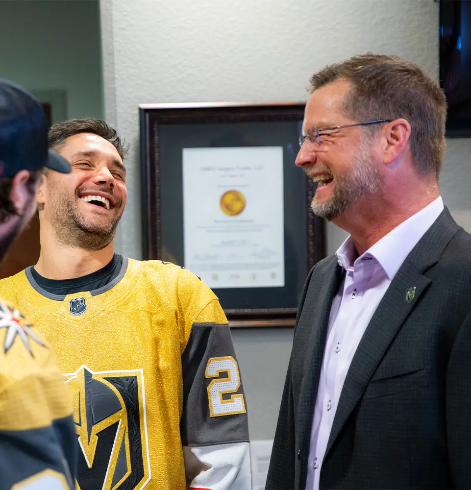Three men are laughing in an office setting. Two wear golden hockey jerseys, while the third is in a suit. A framed certificate is visible in the background.