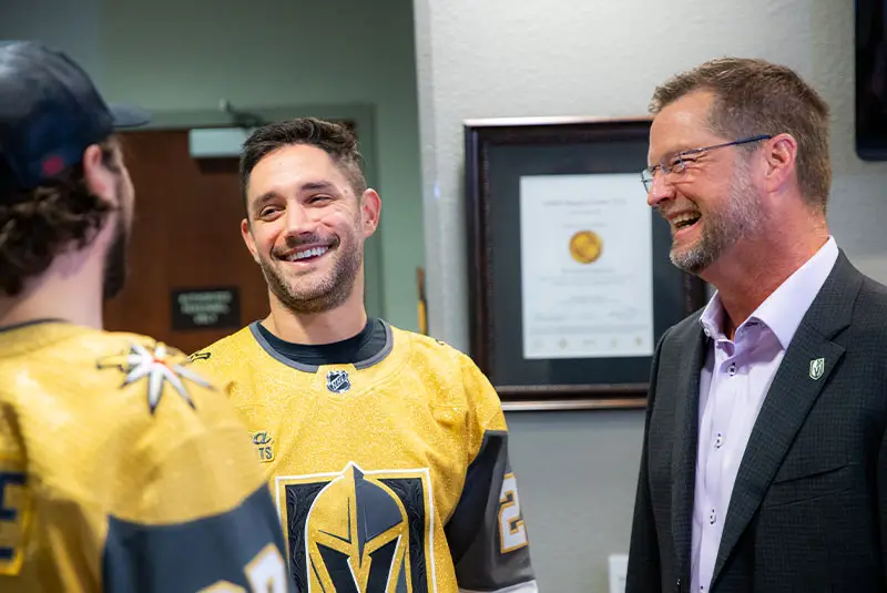 Three men are laughing in an office setting. Two wear golden hockey jerseys, while the third is in a suit. A framed certificate is visible in the background.